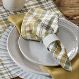 Sage & Mustard Plaid Cloth Napkin in white, sage green, putty brown and mustard yellow is wrapped around a white ribbed napkin ring, placed on a white plate over a larger charger plate, with a yellow napkin and a plaid placemat visible on a rustic wooden table.