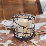 A Barbed Wire Rustic Pillar Candle Holderwith bubble glass wrapped with dark metal barbed wire, containing a lit cream-colored candle, set on a rustic wooden surface with a cowhide rug and blurred wooden background.