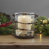 Barbed Wire Rustic Pillar Candle Holder and a lit candle on a wooden surface with Christmas accents.