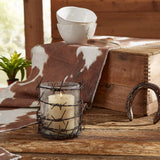 Barbed Wire Rustic Pillar Candle Holder with a barbed-wire cage and clear glass, holding a lit cream candle, styled on a wooden table with a cowhide throw, stacked white bowls, a wooden chest, and a horseshoe.