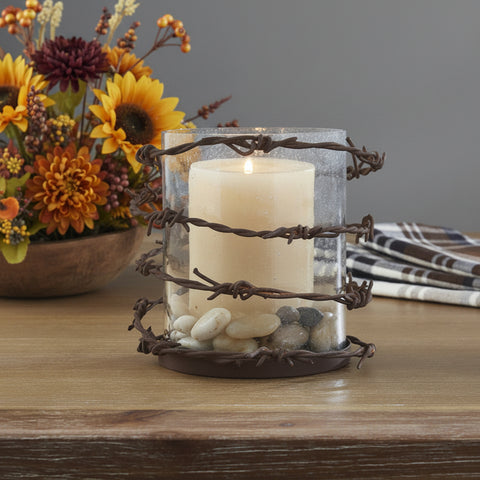 Barbed Wire Pillar Candle Rustic Holder featuring a clear glass cylinder wrapped with dark brown barbed wire, containing a lit cream pillar candle and small decorative stones, set on a wooden tabletop with fall florals
