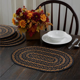 Blackstone Farm Braided Oval Placemat Set of 6 – 12x18 – Black and Tan Jute
 on a dark wood dining table, styled with a white bowl and plate, silverware, a stack of matching placemats, and an autumn floral arrangement with a wooden chair.