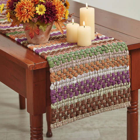 Fall Colors woven Chindi Table Runner with horizontal stripes of green, purple, orange, brown, beige, and white, draped over a dark wood table with Fall florals and candles. 