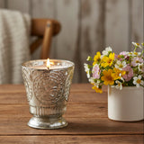 Fleur de Lys Silver Tumbler Hand-Poured Soy Scented Candle - a silver-toned glass candle holder with embossed floral detailing sits on a rustic wooden table next to a small white vase of yellow, pink, and white flowers.