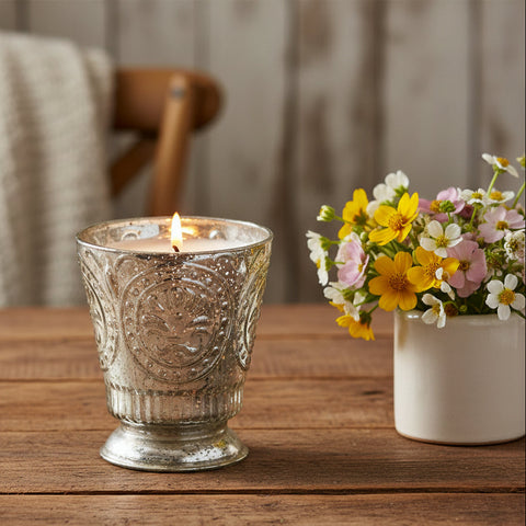 Fleur de Lys Silver Tumbler Hand-Poured Soy Scented Candle - a silver-toned glass candle holder with embossed floral detailing sits on a rustic wooden table next to a small white vase of yellow, pink, and white flowers.