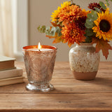 Fleur de Lys Tangerine Tumbler Hand-Poured Soy Scented Candle in a textured, footed glass holder with a distressed silver and copper-orange finish, set on a table with Fall florals and a stack of books.