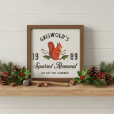 Griswold's Squirrel Removal Holiday Sign 6x6 with a distressed brown wood frame on a wood shelf with Christmas holly and pinecones. The sign features a vintage-style illustration of a red squirrel holding a nut with green leaves and red berries, set against an off-white background.