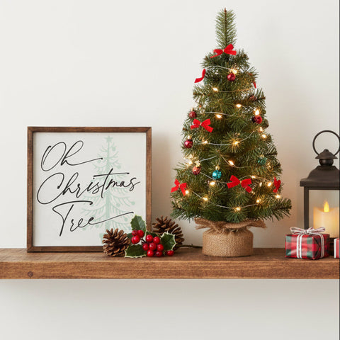 Oh Christmas Tree Holiday Sign 10x10 A small decorated Christmas tree stands on a wooden shelf beside a framed sign reading 'Oh Christmas Tree,' with pinecones, holly, a lantern, and wrapped gifts in a warm holiday setting.