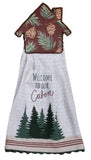 A cabin house-shaped kitchen towel with a brown pine tree print on the top half and a white bottom half that reads 'Welcome to our Cabin' above green pine trees, shown on a white background.