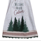A house-shaped kitchen towel with a brown pine tree print on the top half and a white bottom half that reads 'Welcome to our Cabin' above green pine trees, shown on a white background.