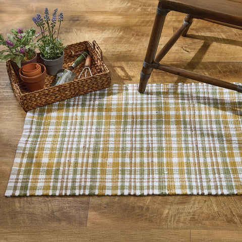 Sage & Mustard Plaid Chindi Rag Rug on a wooden floor with a basket of plants.