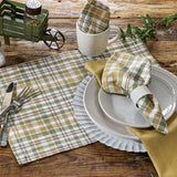 Sage & Mustard Plaid Cloth Napkin in white, sage green, putty brown and mustard yellow is wrapped around a white ribbed napkin ring, placed on a white plate over a larger charger plate, with a yellow napkin and a plaid placemat visible on a rustic wooden table with a white mug that contains the Sage & Mustard Dishcloth.