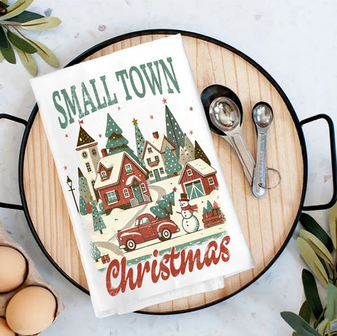 A folded white kitchen towel with a festive Christmas illustration and the words 'SMALL TOWN Christmas' is displayed on a round wooden serving tray. The towel is styled with metal measuring cups, eggs, and greenery on a light marble surface.