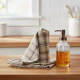 Tinderbox Brown Plaid Dishcloths stacked on a wood counter beside a white farmhouse sink, glass soap dispenser, and potted herbs — set of 3 waffle weave cotton dishcloths in brown and oatmeal plaid