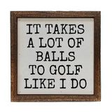 A square framed decorative sign with a rustic brown wood frame and a white face. The sign features the humorous text 'IT TAKES A LOT OF BALLS TO GOLF LIKE I DO' in a black, hand-drawn serif font.